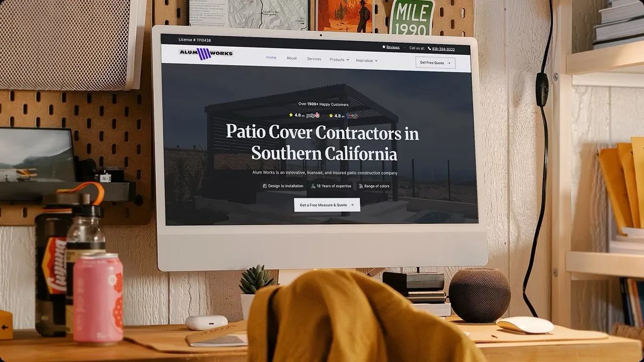 The Alum Works homepage on an iMac at a timber desk in front of a pegboard wall, its hero reading "Patio Cover Contractors in Southern California" over a dusk shot of a black louvred pergola, with Yelp and Google ratings above.