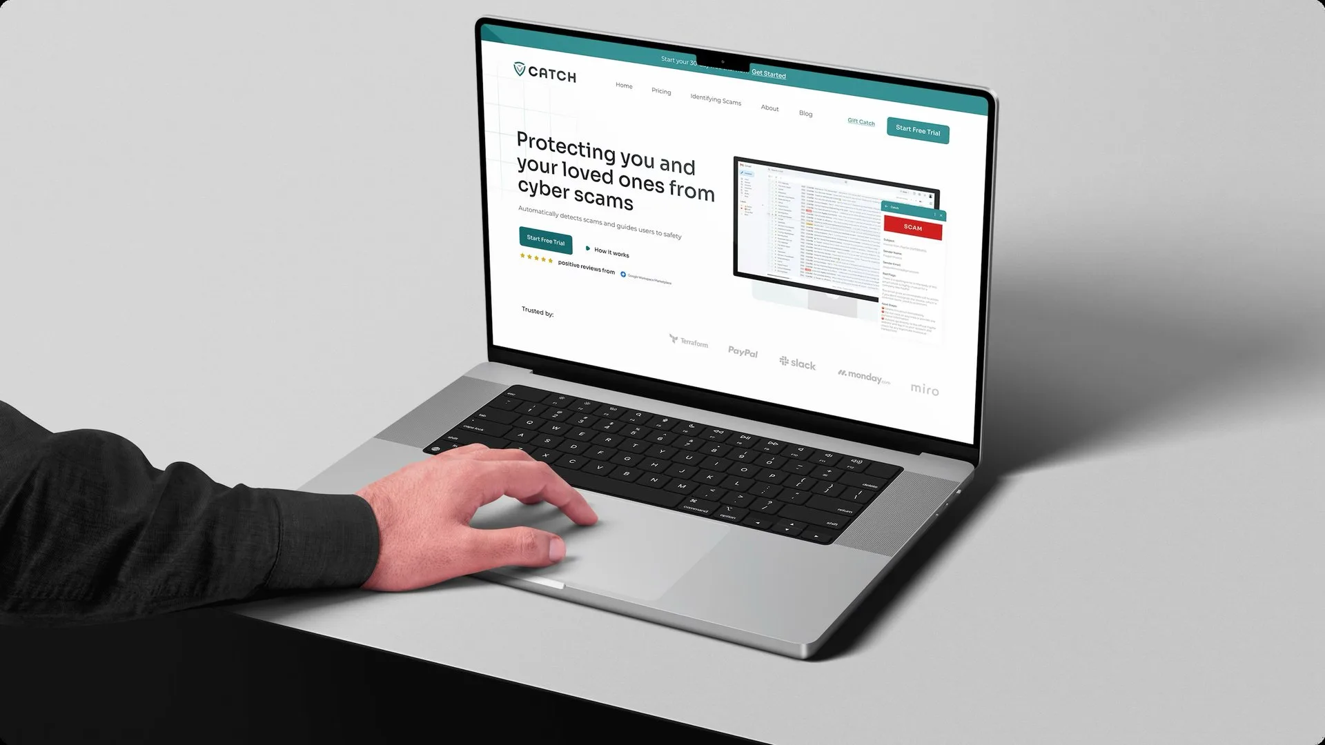 The Catch homepage open on a MacBook angled on a pale grey desk with a hand resting on the trackpad, showing the Protecting you and your loved ones from cyber scams headline beside a Gmail inbox flagged with a red SCAM warning.