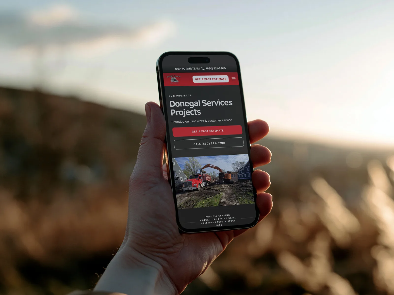 The Donegal Services projects page on an iPhone held up outdoors at sunset, headed "Donegal Services Projects" above a photo of a dump truck and excavator on a cleared site.