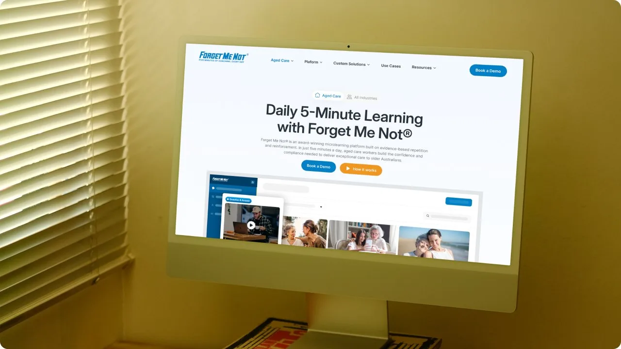 The Forget Me Not homepage on an iMac in a warm-lit room, headed “Daily 5-Minute Learning with Forget Me Not” above the aged-care microlearning interface.