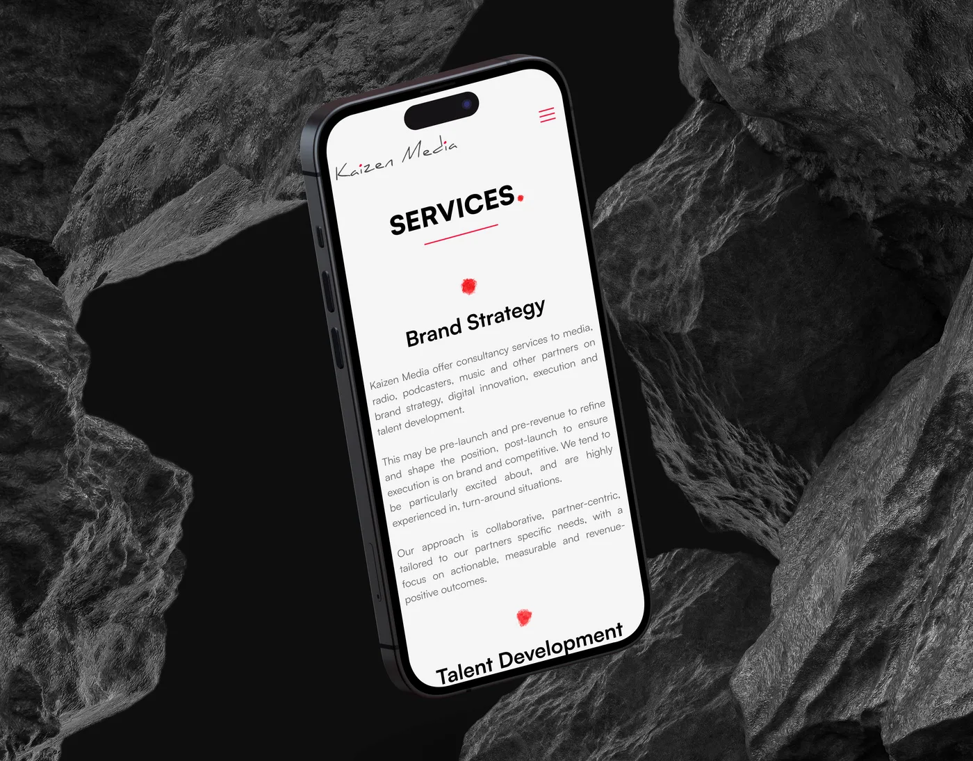 The Kaizen Media Services page on an iPhone angled among dark volcanic rocks, listing Brand Strategy and Talent Development.