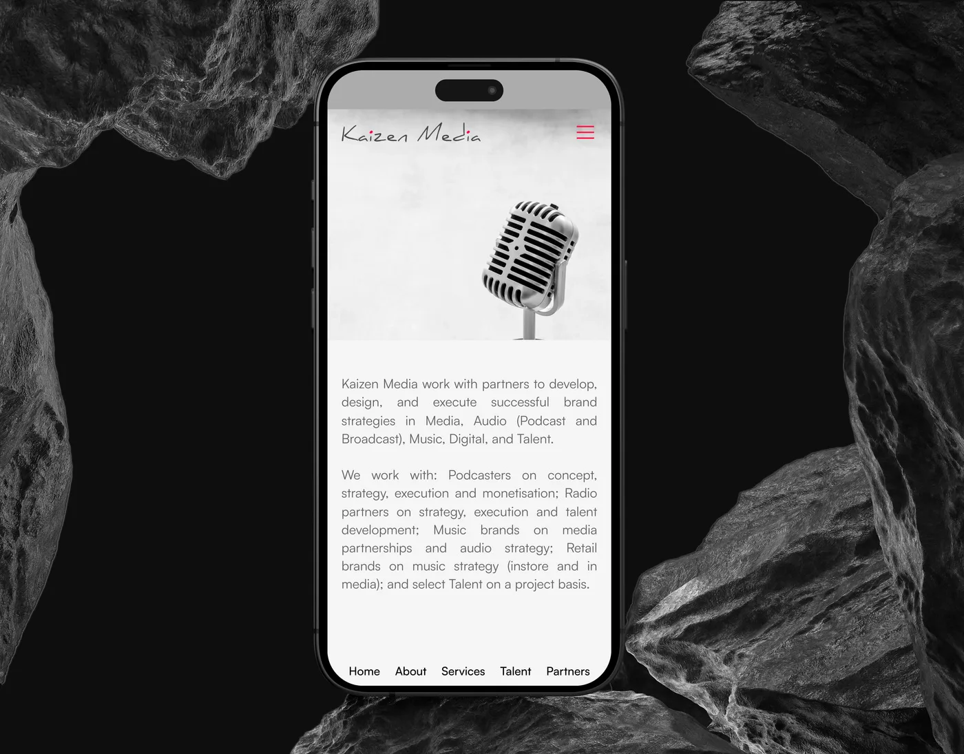 The Kaizen Media mobile homepage on an iPhone set upright between dark rocks: a vintage microphone above the consultancy’s introduction.
