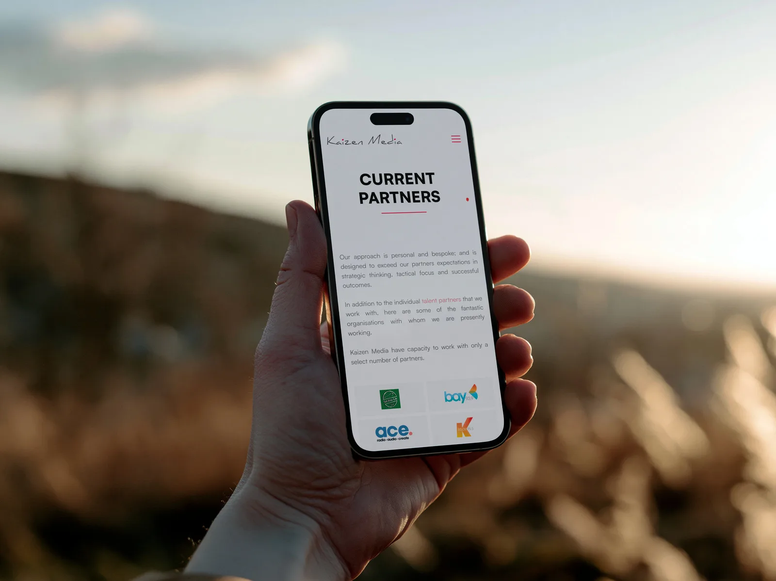 The Kaizen Media Current Partners page on an iPhone held up outdoors at sunset, with partner logos below the introduction.
