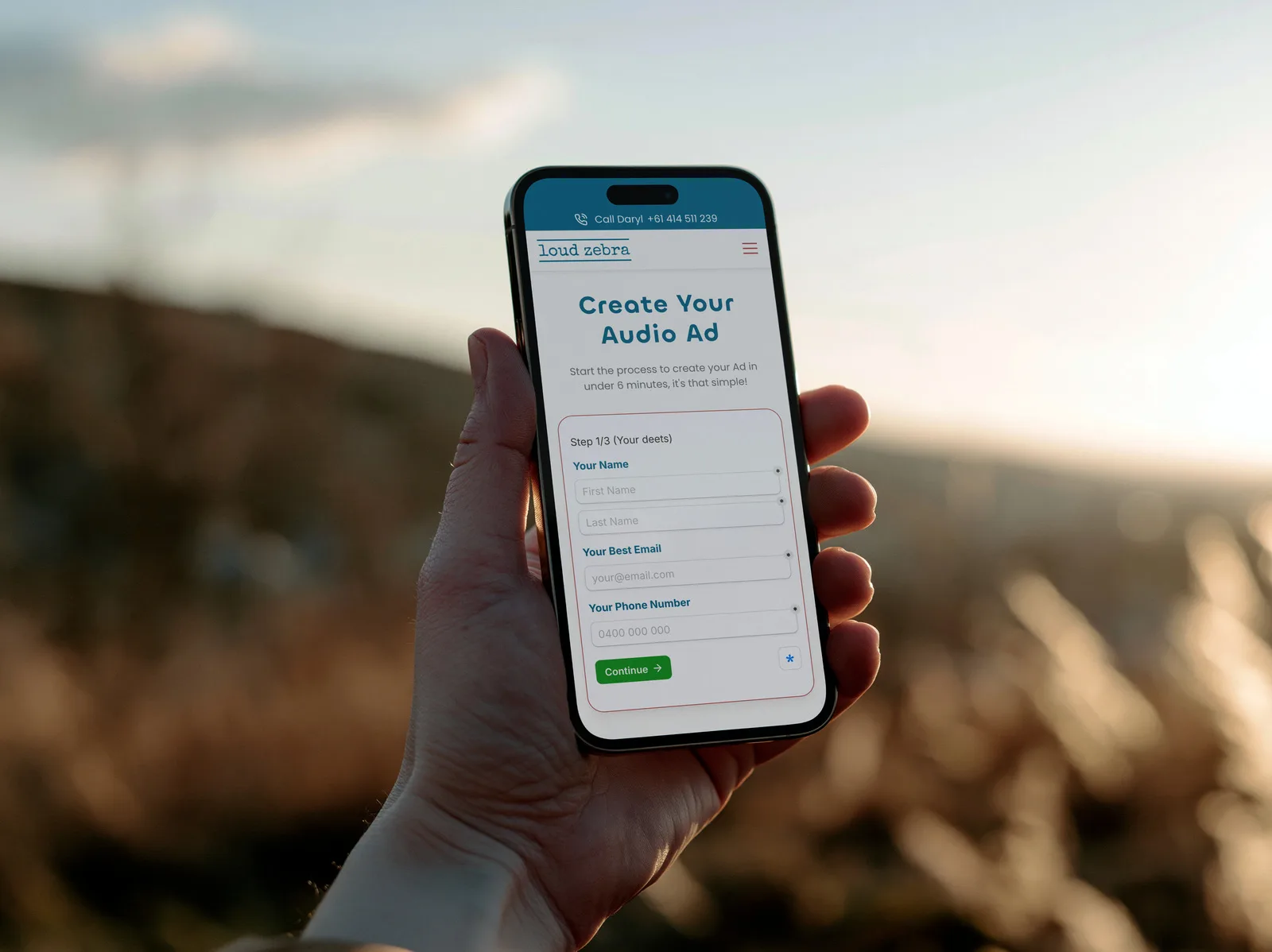 The Loud Zebra Create Your Audio Ad form on an iPhone held up outdoors at sunset, open at step 1 of 3.