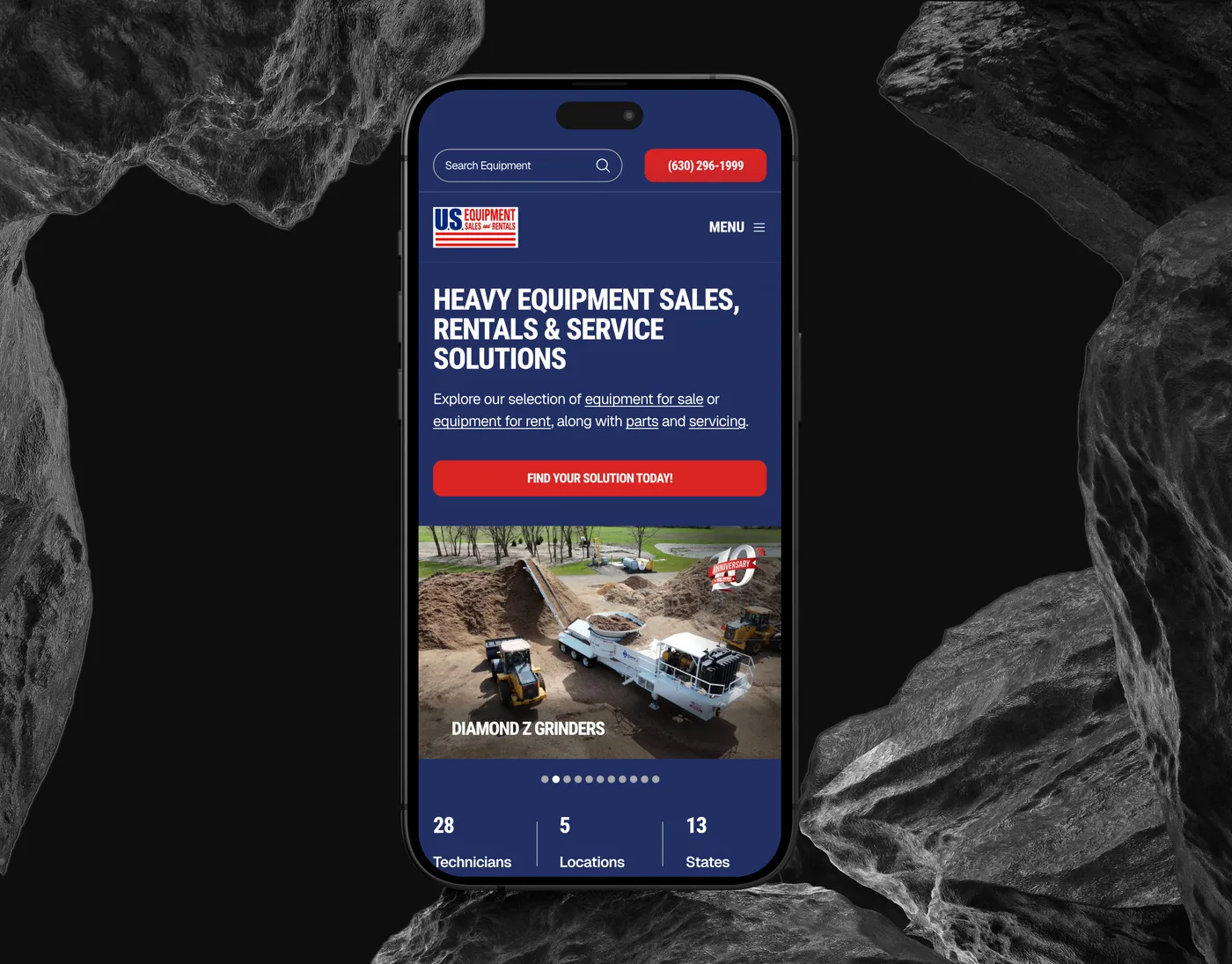 The US Equipment Sales mobile homepage on an iPhone set upright between dark rocks, headed "Heavy Equipment Sales, Rentals & Service Solutions" above a photo of a grinder at work.